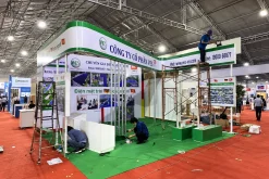 Nhân sự đang lắp đặt gian hàng có thảm xanh, quầy trắng và khung bảng hiệu phía trên.
