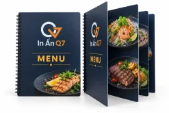 Menu nhựa PVC đóng lò xo màu navy và cam In Quảng Cáo Q7, gồm cuốn đóng và cuốn mở trên nền trắng.