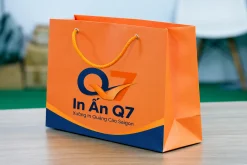 Túi giấy màu cam in nhận diện In Quảng Cáo Q7, quai dây màu vàng.