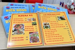 Menu hải sản mở hai trang màu vàng, phía sau là các cuốn menu có bìa xanh.
