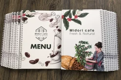 Các cuốn menu cà phê có bìa in hình lá và hạt cà phê, đóng lò xo trắng.